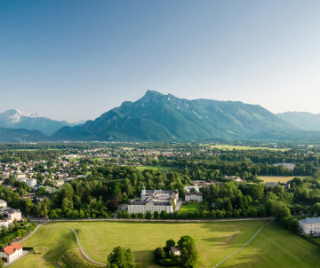 Panoramic view across Salzburg, Austria, with residential neighbourhoods, green parkland and historic buildings extending toward Untersberg Mountain. Forested foothills and Alpine peaks define the skyline in the distance.