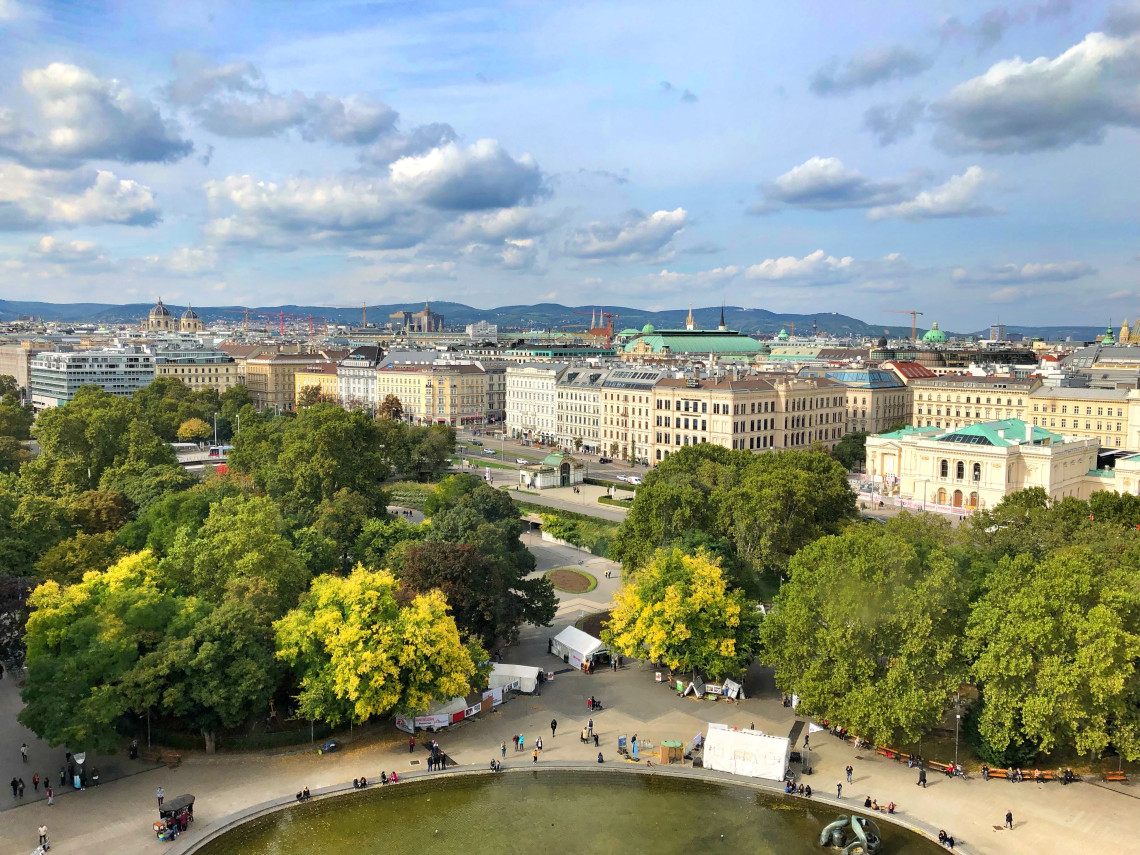 Elevated view across Stadtpark in Vienna, Austria, with mature trees, walking paths and a pond surrounded by historic buildings. The city centre extends toward low hills beneath a partly cloudy sky.