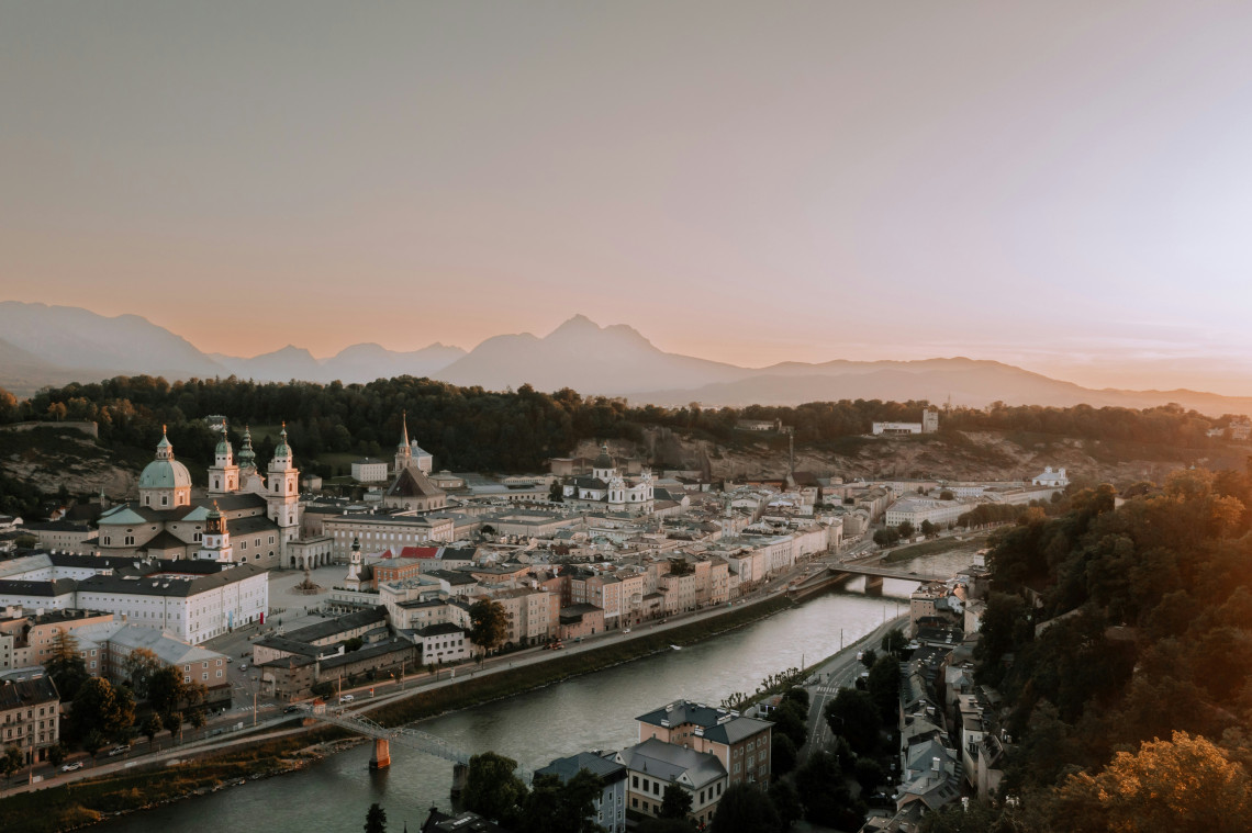 Panoramic view of Salzburg Old Town in Salzburg, Austria, with the Salzach River running past church domes, historic buildings and tree-covered hills. Alpine peaks appear in the distance beneath warm evening ligh