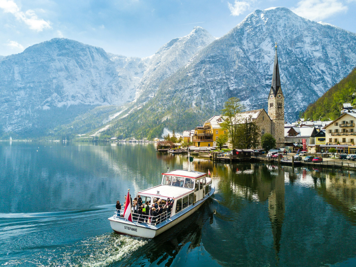 A passenger boat travels across Lake Hallstatt beside the village of Hallstatt, Austria. The lakeside church, waterfront buildings and steep Alpine mountains are reflected in the calm water.