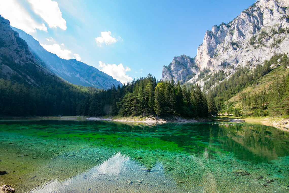 Gr&uuml;ner See in Trag&ouml;&szlig;, Austria, features transparent green water bordered by dense forest and limestone mountains. The shallow Alpine lake reveals stones and aquatic vegetation beneath its surface.