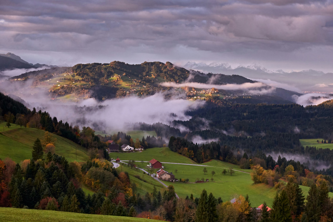 Mist drifts between forested hills and green farmland in the Styrian countryside of Austria. Scattered farmhouses, autumn trees and distant mountain ridges extend beneath a cloudy sky.
