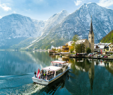 A passenger boat crosses Lake Hallstatt in Hallstatt, Austria, with the lakeside village and its church reflected in the calm water. Steep Alpine mountains rise behind the settlement in the Salzkammergut region.