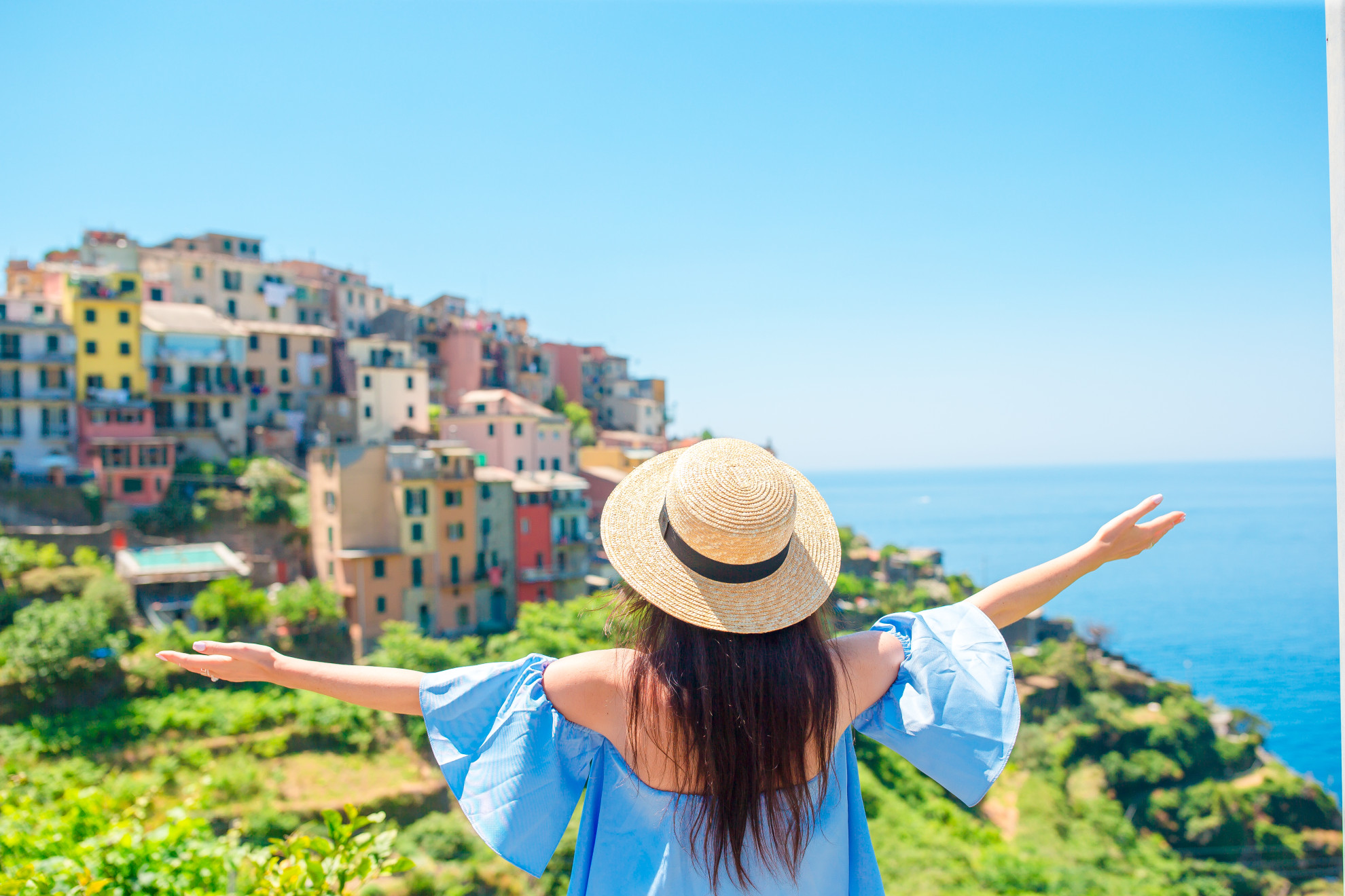 Woman with straw hat and arms wide open overlooking the colorful cliffside village of Corniglia in Cinque Terre, Italy