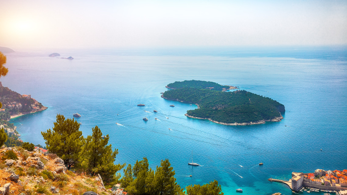 Panoramic view of forested Lokrum Island off Dubrovnik, Croatia, surrounded by clear Adriatic waters with boats sailing between the island and the historic city.