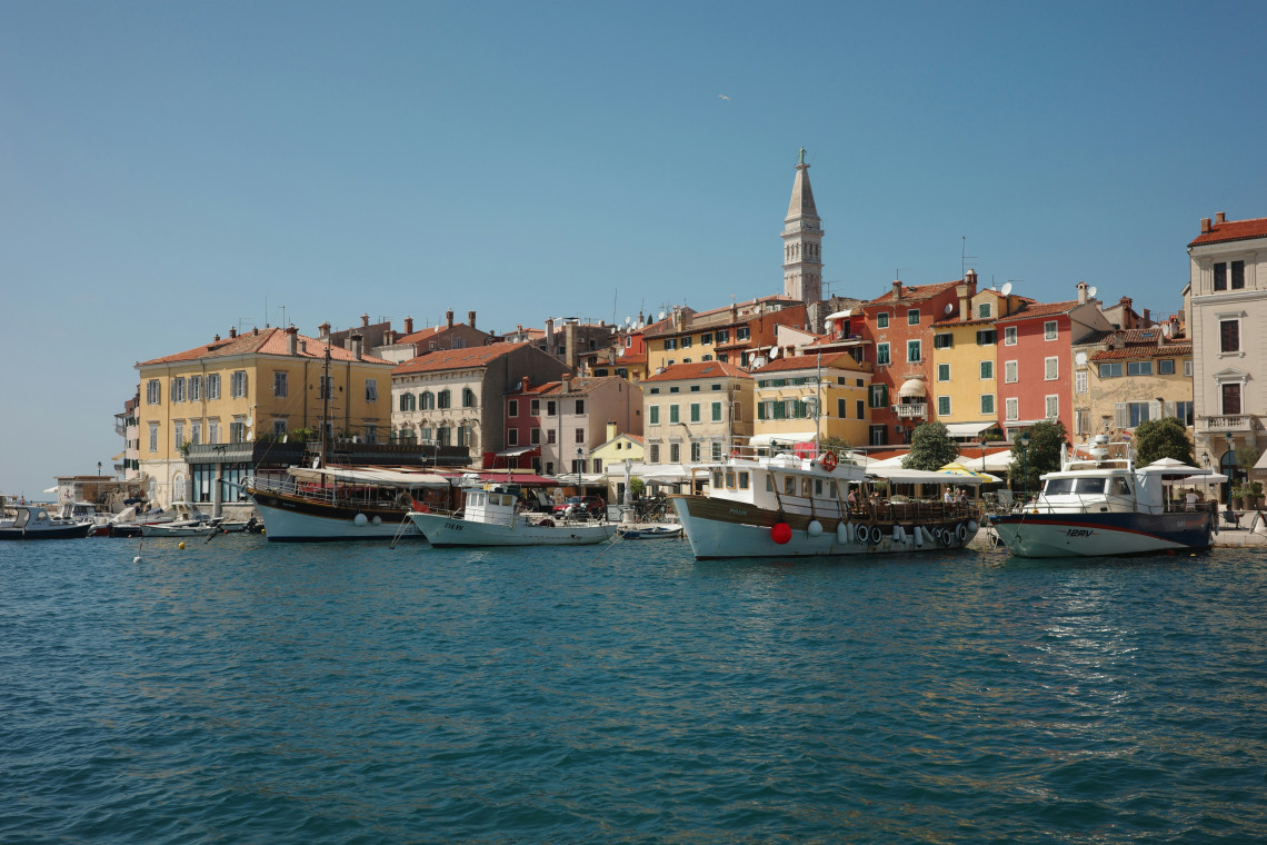Boats moored along the waterfront of Rovinj, Croatia, with colourful historic buildings and the bell tower of the Church of St Euphemia rising above the Old Town.