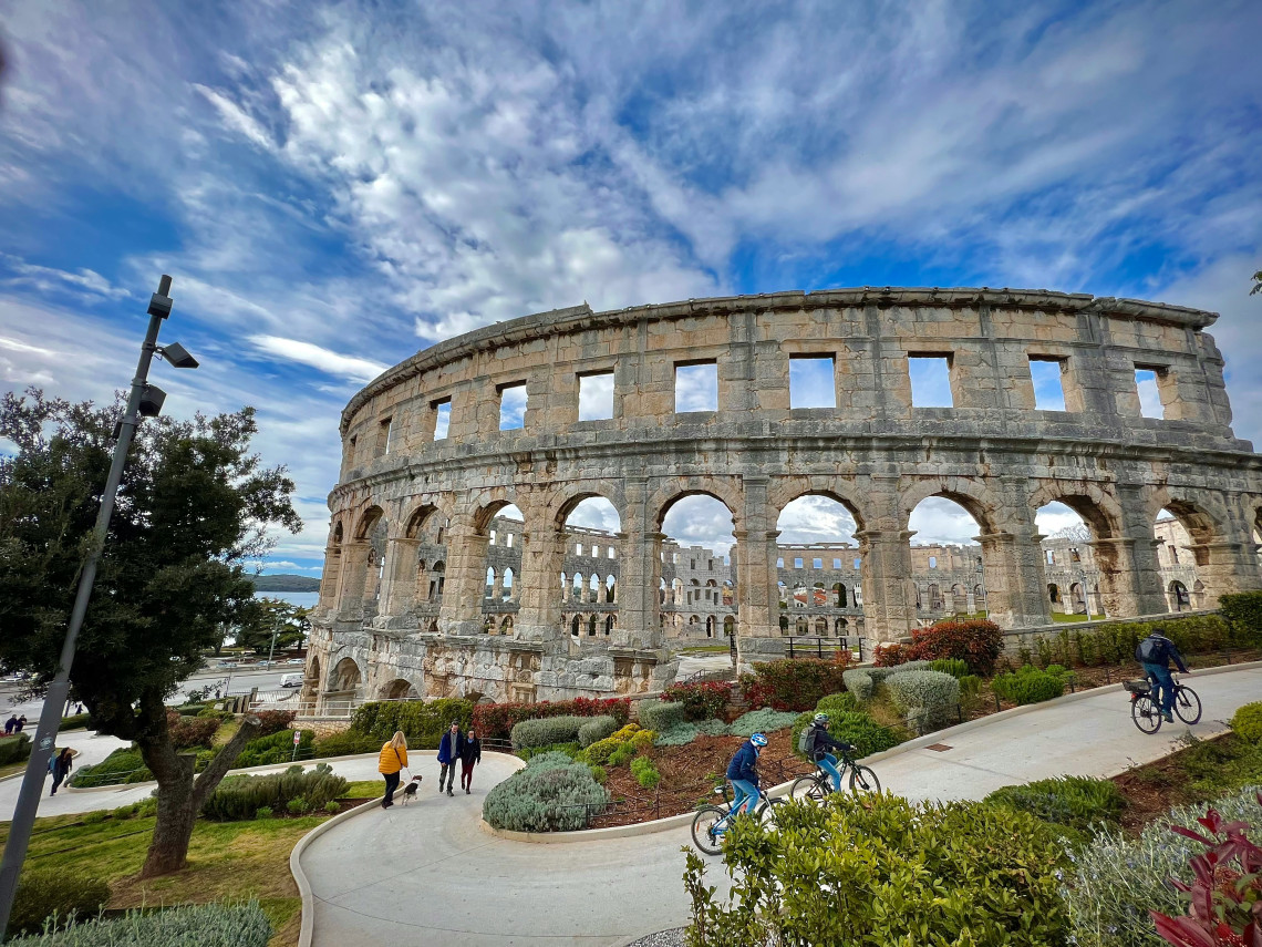 Exterior view of the Pula Arena in Pula, Croatia, showing the preserved stone arches of the ancient Roman amphitheatre surrounded by landscaped paths and Mediterranean vegetation.