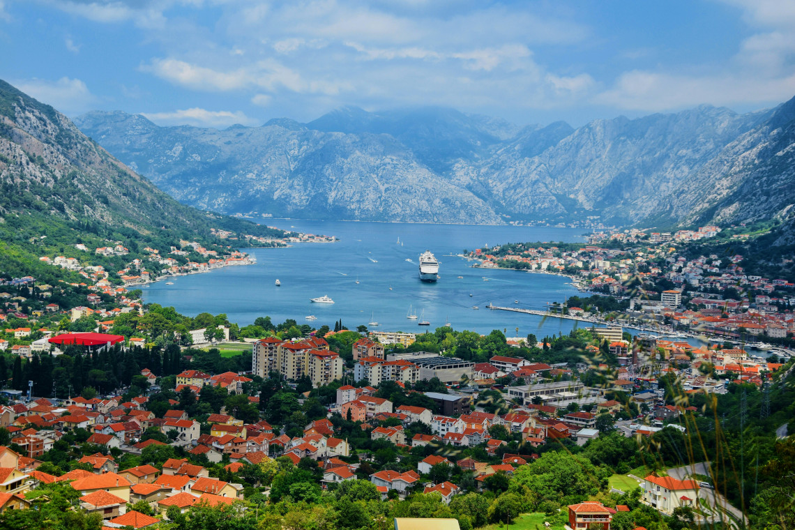 Panoramic view across the Bay of Kotor in Montenegro, where a cruise ship sails between waterfront settlements beneath steep limestone mountains