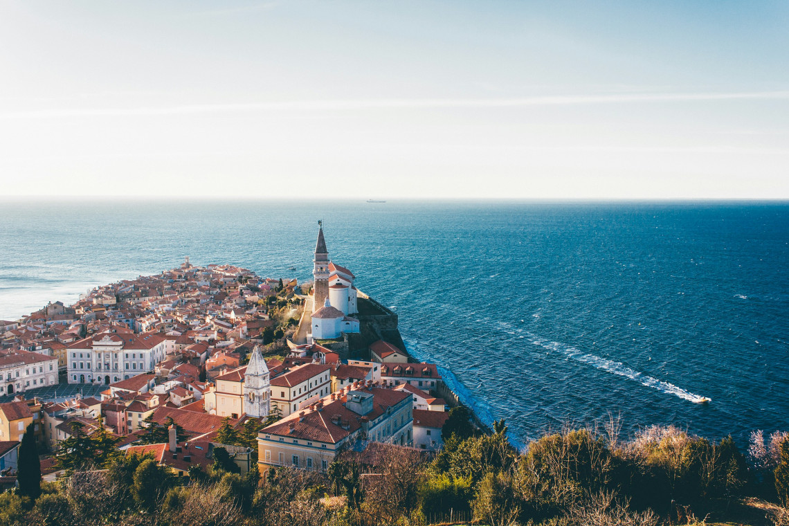 Elevated view of Piran, Slovenia, with the Church of St George rising above terracotta rooftops on a narrow peninsula extending into the Adriatic Sea.