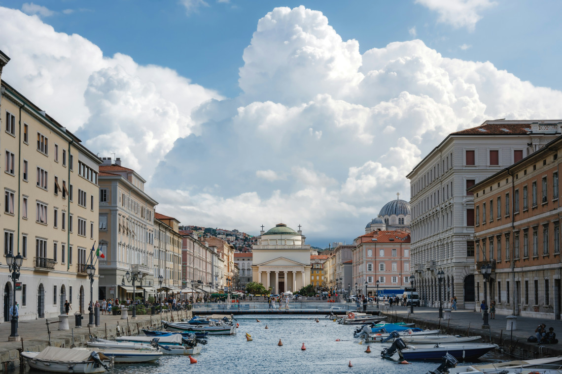 View along the Grand Canal in Trieste, Italy, lined with historic buildings and small boats, with the neoclassical Church of Sant&rsquo;Antonio Taumaturgo at the end of the canal and the dome of Saint Spyridon Church visible nearby.