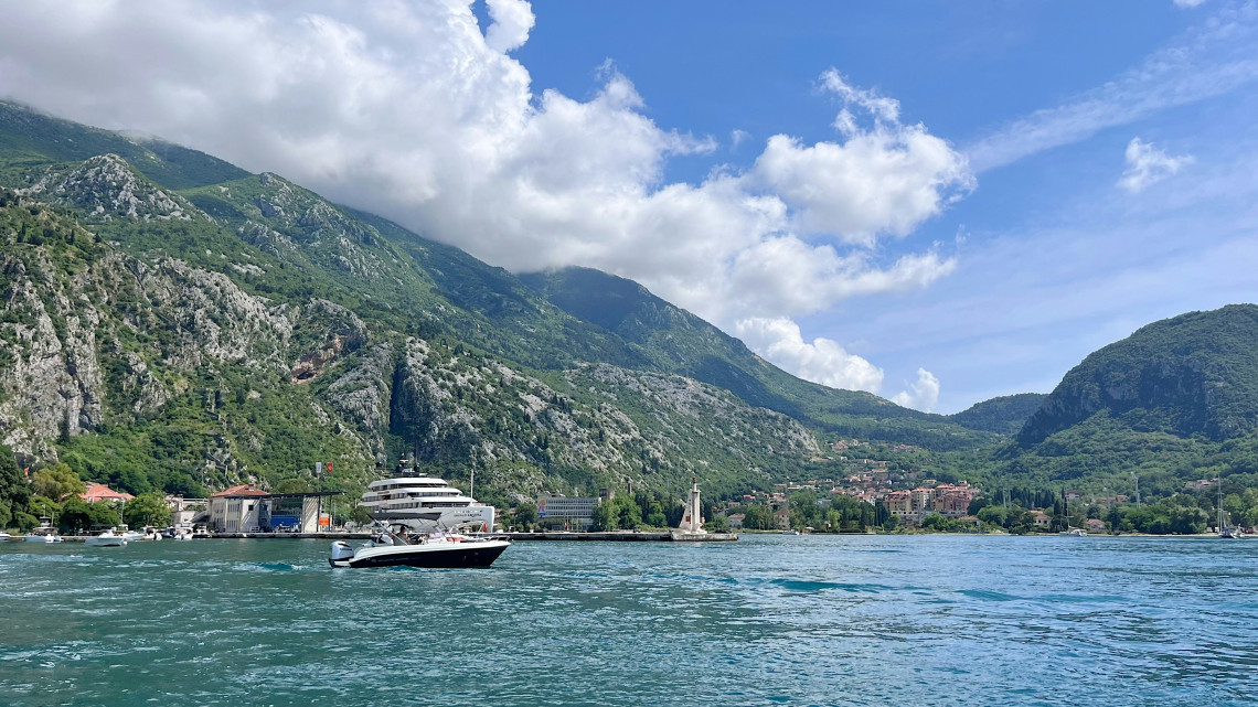 View across the Bay of Kotor in Montenegro, with boats crossing calm blue water and dramatic limestone mountains rising directly behind the historic coastal settlements.