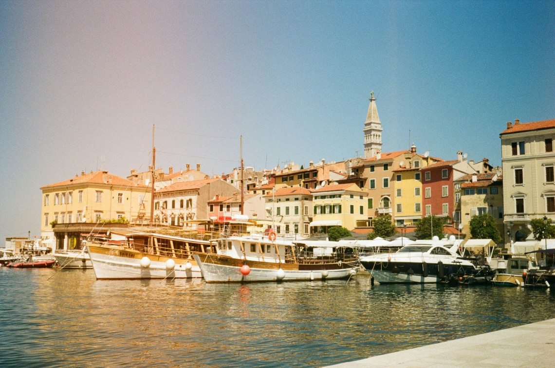 Traditional boats moored along Rovinj Harbour in Croatia, backed by colourful historic buildings and the bell tower of the Church of St Euphemia in the Old Town.