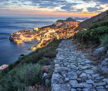 Elevated view of Dubrovnik, Croatia, glowing along the Adriatic coast at dusk, with the historic Old Town and surrounding coastline seen from a rocky hillside path.