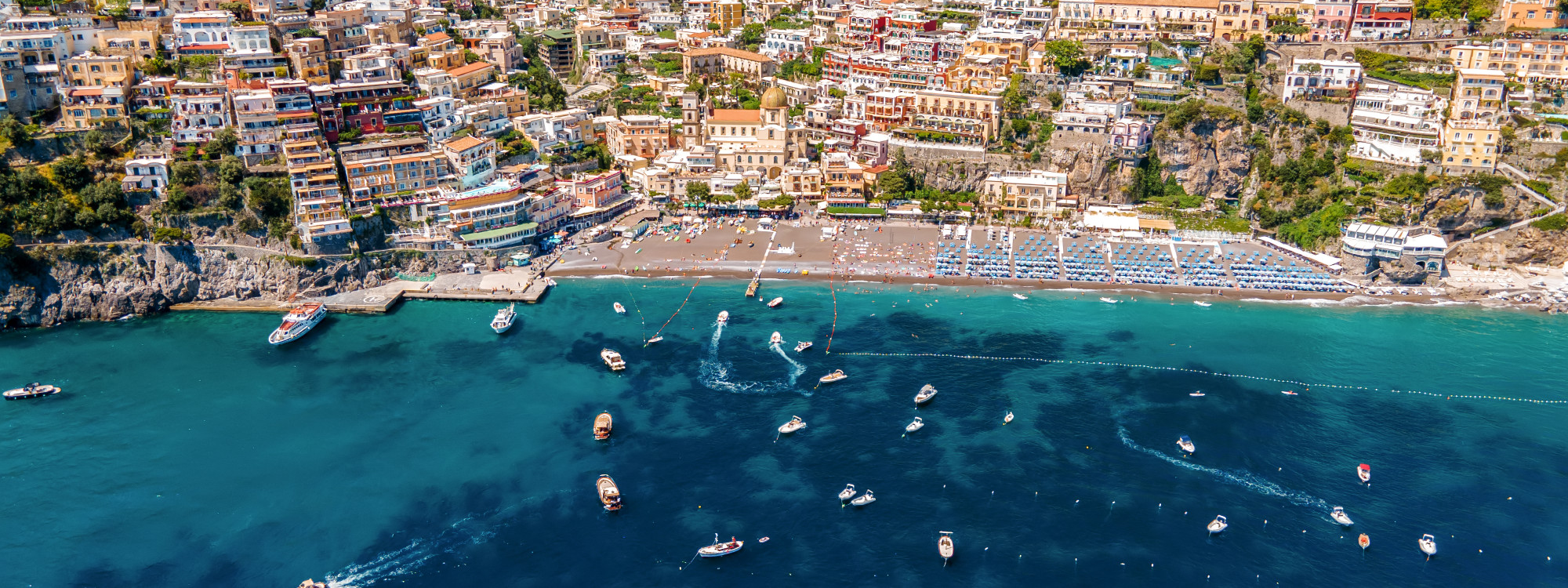 Aerial view of Marina Grande Beach in Positano on Italy’s Amalfi Coast. Beach facilities line the shoreline while boats float in the turquoise water below the densely built hillside town.