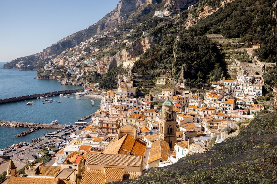 Elevated view over Amalfi on Italy&rsquo;s Amalfi Coast, with the bell tower of Amalfi Cathedral rising among terracotta rooftops. The town descends toward its harbour beneath steep, terraced coastal mountains.
