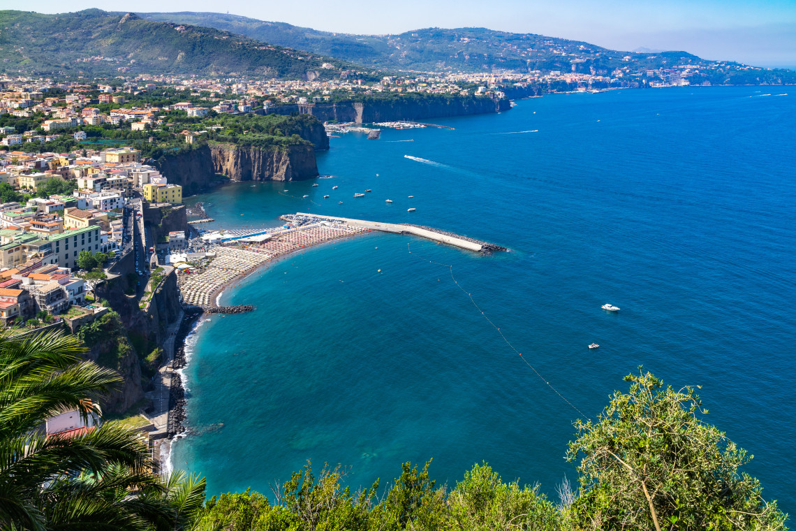 Elevated view of the beach at Meta on the Sorrento Peninsula in Campania, Italy. The broad shoreline and protective breakwater curve beneath steep coastal cliffs, with the Gulf of Naples extending into the distance.