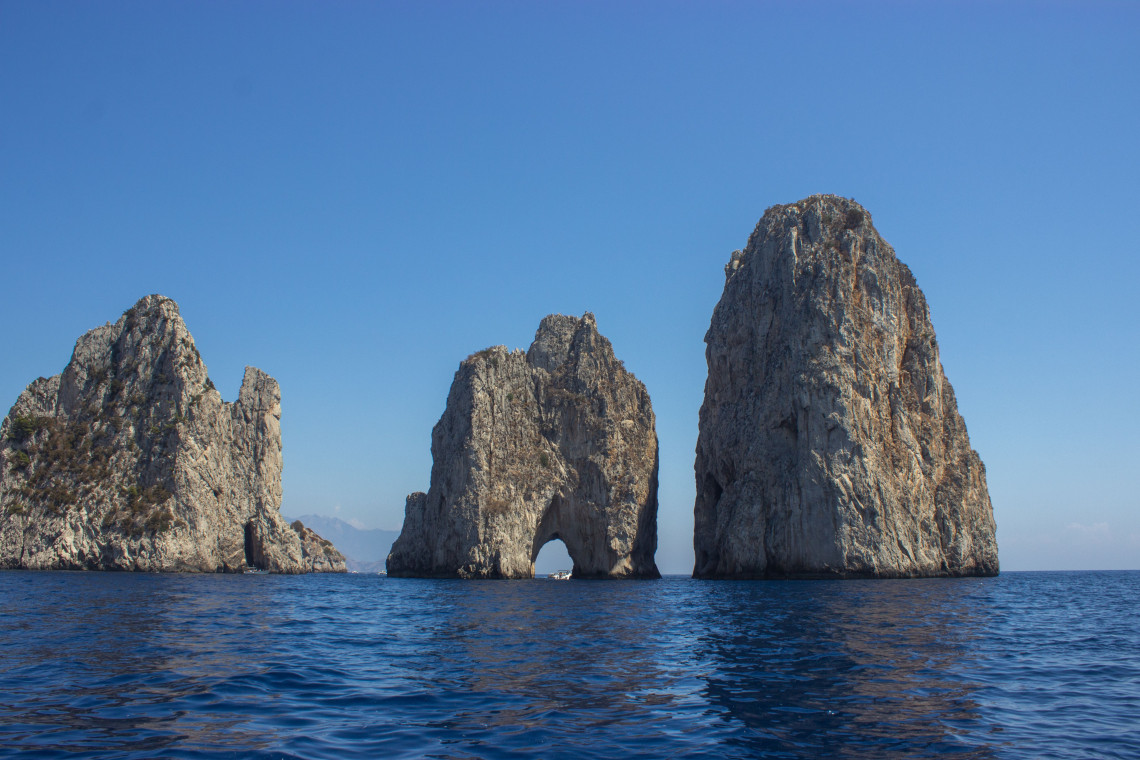 The Faraglioni rock formations rise from the Mediterranean Sea off Capri, Italy. Three towering limestone stacks dominate the open water beneath a clear blue sky, including the central rock with its natural arch.