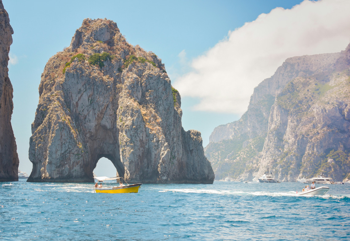 Boats cruise near Faraglione di Mezzo off the coast of Capri, Italy, with its large natural arch visible at sea level. Steep limestone cliffs and deep blue Mediterranean water surround the landmark.
