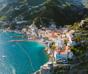Aerial view of Amalfi on Italy’s Amalfi Coast, showing the historic town clustered between steep mountains and the Mediterranean Sea. The marina, waterfront buildings and cathedral stand beside clear blue coastal waters.