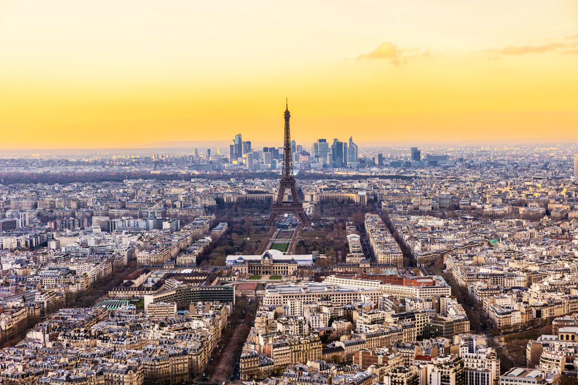 Panoramic view across Paris with the Eiffel Tower rising above the city skyline in warm sunset light and La D&eacute;fense visible in the distance.