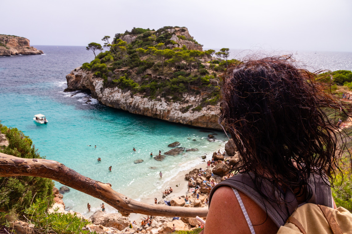 Visitor overlooking a secluded turquoise cove in Spain&rsquo;s Balearic Islands, with a small sandy beach, limestone cliffs and pine-covered Mediterranean landscape.