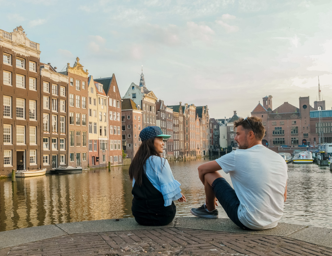 Couple sitting beside the water at Damrak in central Amsterdam, with narrow historic canal houses and boats lining the waterfront.