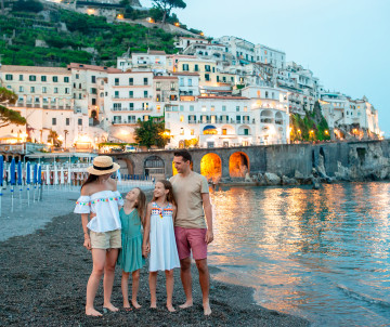 Family standing on the beach in Amalfi, Italy, with the historic hillside town and its white Mediterranean buildings rising above the waterfront at dusk.