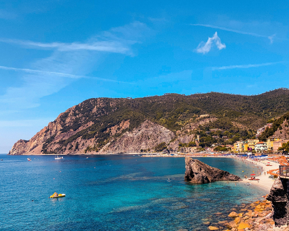 Wide view of the beach at Monterosso al Mare in Cinque Terre, Italy, with clear turquoise water, a rocky outcrop and rows of beach umbrellas. Green hills rise behind the coastal village.