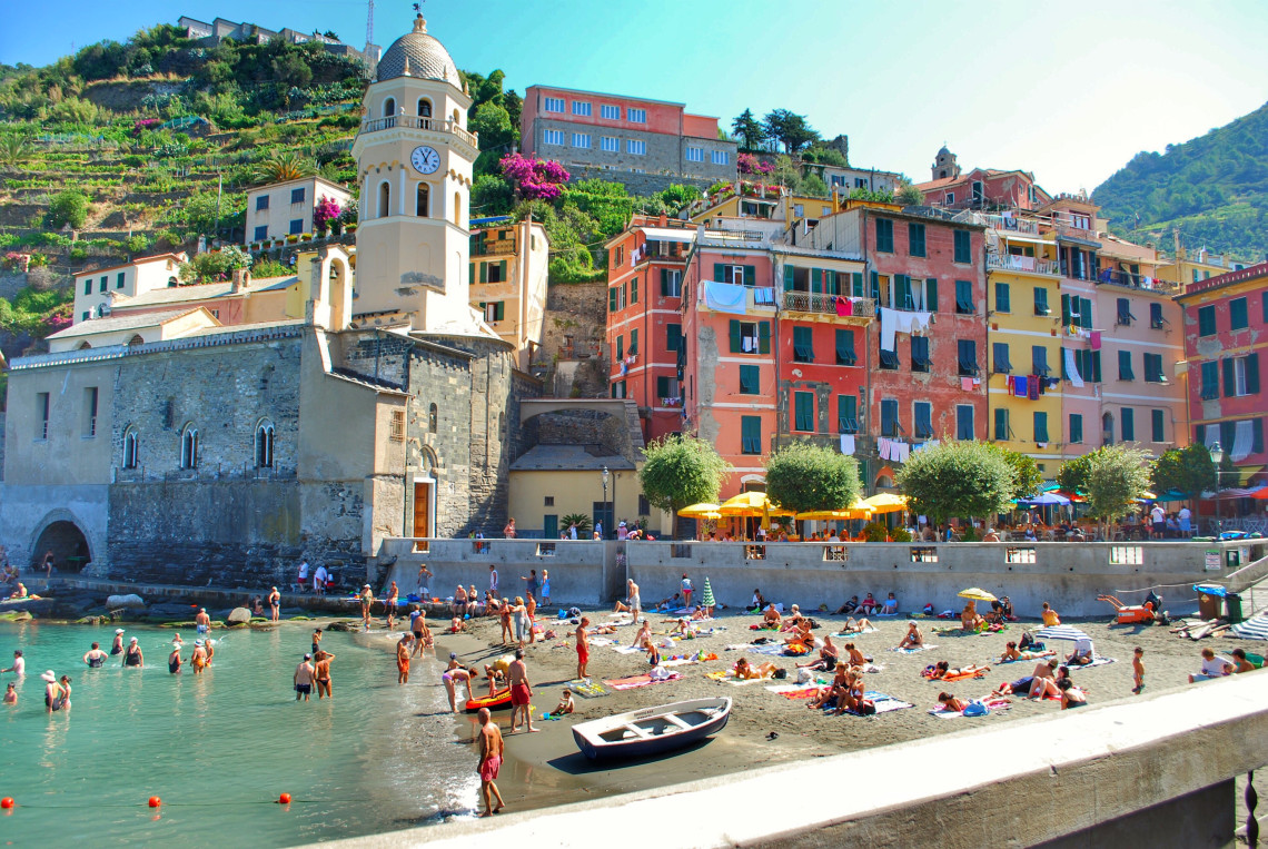 Vernazza Harbour in Cinque Terre, Italy, with visitors gathered along the small beach beneath colourful hillside houses. The Church of Santa Margherita di Antiochia and its bell tower stand directly beside the Ligurian Sea.