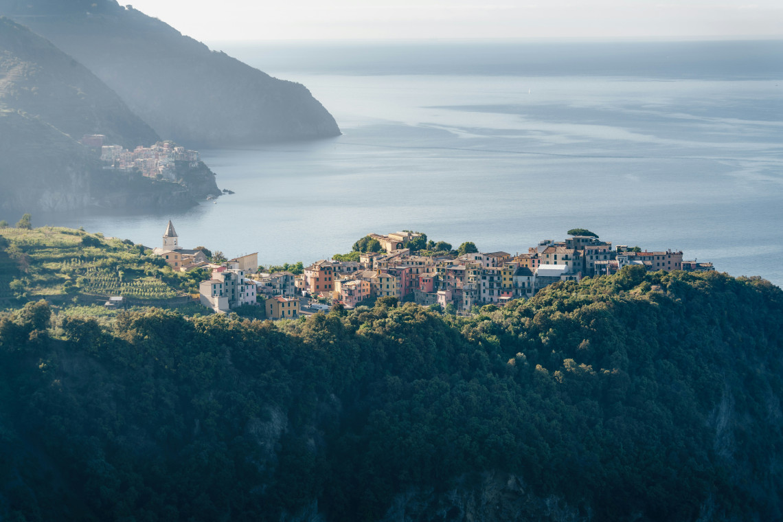 Corniglia sits on an elevated ridge above the Ligurian Sea in Cinque Terre, Italy. Colourful houses and terraced vineyards occupy the green coastal landscape, with another Cinque Terre village visible in the distance.