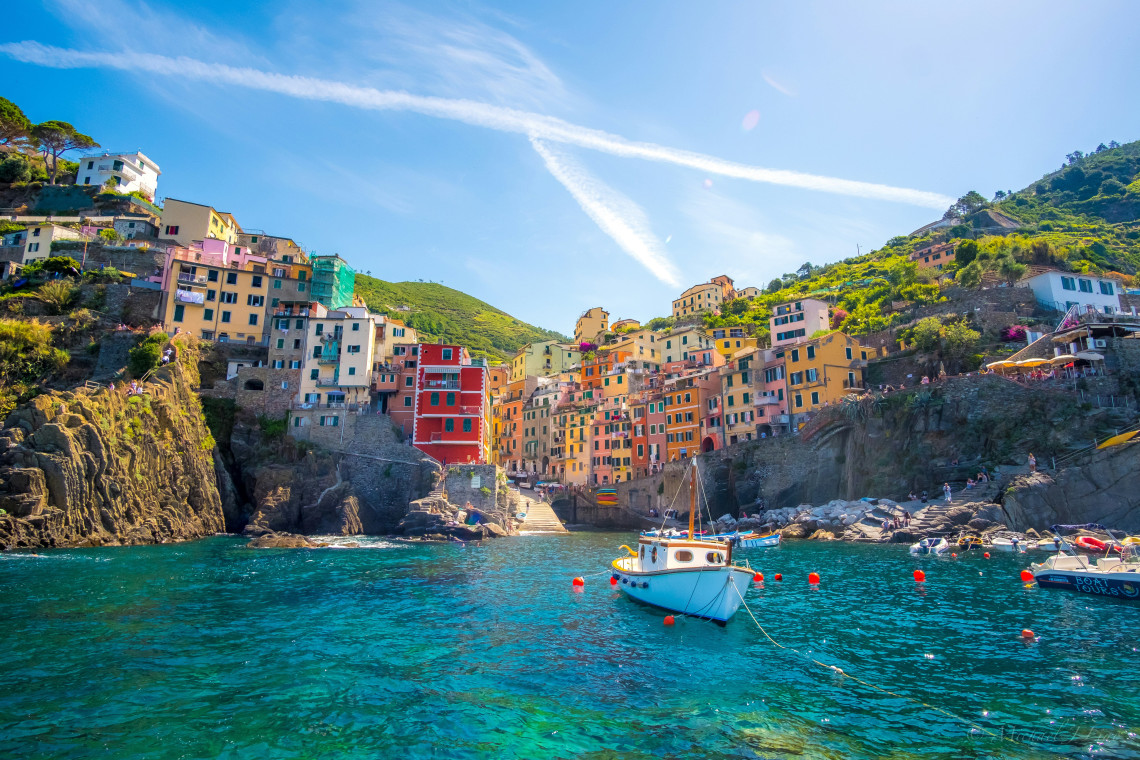 A fishing boat floats in the harbour at Riomaggiore in Cinque Terre, Italy. Tall pastel-coloured houses rise between steep rocky cliffs above the clear blue Ligurian Sea.