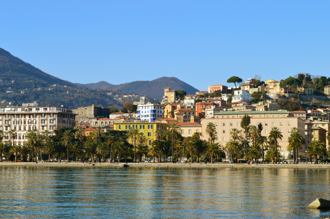 The waterfront of La Spezia, Italy, stretches beside calm waters with palm trees lining Passeggiata Morin. Colourful buildings and green hills rise behind the seafront promenade.