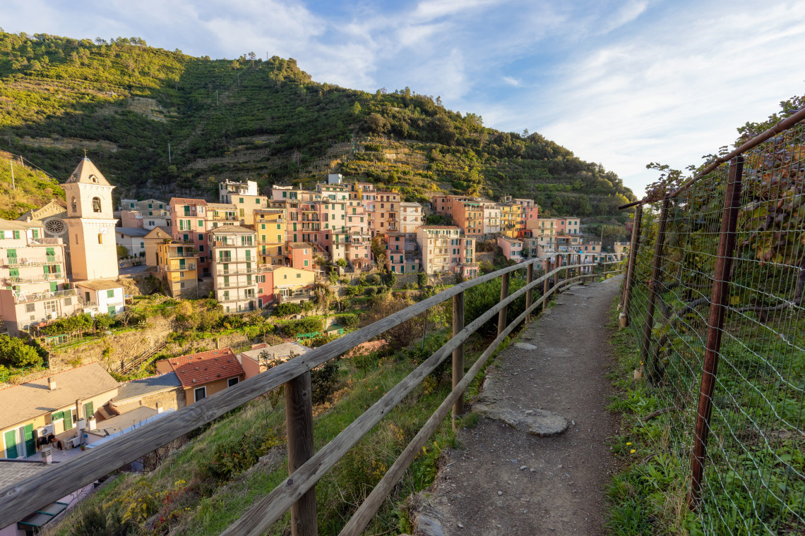 A narrow hiking path follows the terraced hillside above Manarola in Cinque Terre, Italy. Colourful village houses and a church tower sit below slopes shaped by traditional vineyards and dry-stone walls.