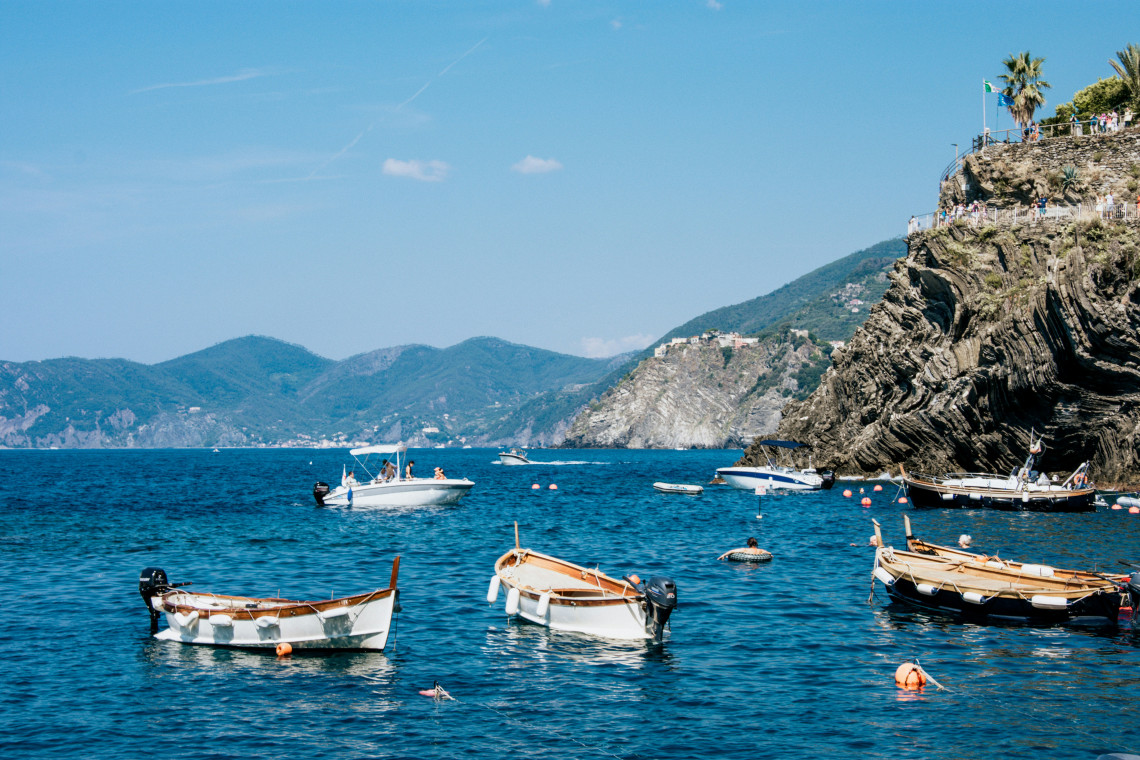 Traditional fishing boats float in the blue waters off Riomaggiore in Cinque Terre, Italy. Steep layered cliffs frame the small harbour against the mountainous Ligurian coastline.
