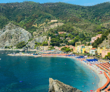 Monterosso Beach curves along the coast of Monterosso al Mare in Cinque Terre, Italy. Clear blue water meets the beach beneath steep green hills and the rocky Ligurian coastline.