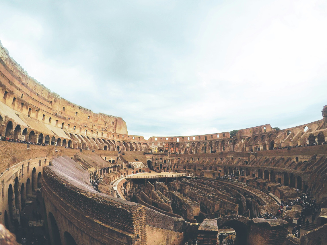 Panoramic interior view of the Colosseum in Rome, Italy, showing the arena, underground chambers and multiple levels of ancient seating. Visitors can be seen exploring the historic Roman amphitheatre.