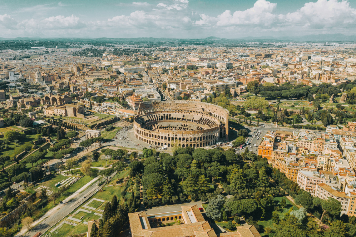 Aerial view of the Colosseum and surrounding archaeological area in central Rome, Italy. The amphitheatre is surrounded by the ruins, green spaces and dense urban fabric of the historic city.
