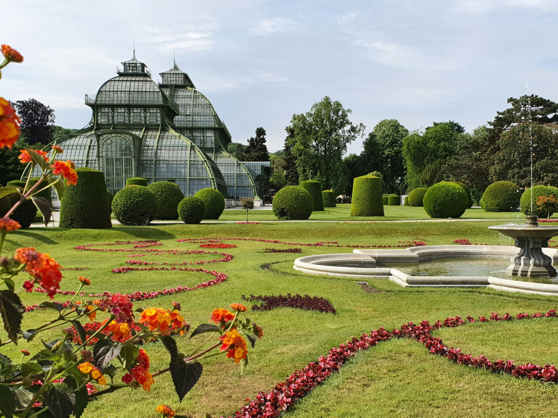 The historic Sch&ouml;nbrunn Palm House in Vienna, surrounded by manicured gardens, sculpted hedges and colorful flower beds.