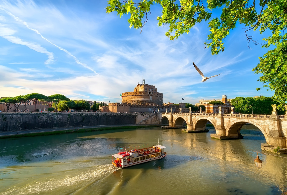 Castel Sant&rsquo;Angelo beside the Tiber River in Rome, with Ponte Sant&rsquo;Angelo crossing the water and a sightseeing boat passing below.