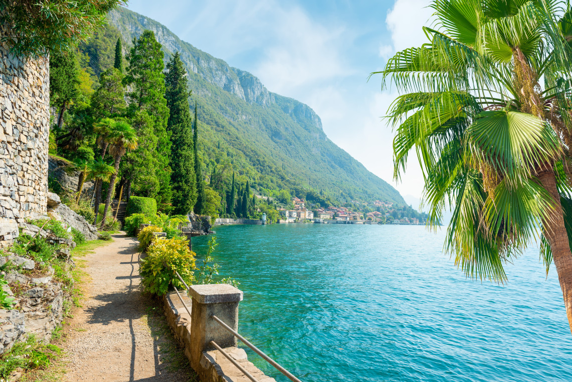 Scenic walking path along Lake Como in Lombardy, with turquoise water, Mediterranean vegetation, steep green mountains and lakeside villages.