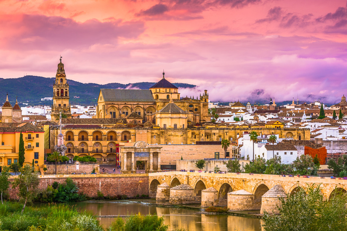 View of the Mosque-Cathedral of C&oacute;rdoba and its bell tower beside the Roman Bridge, with the Guadalquivir River and historic city center at sunset.