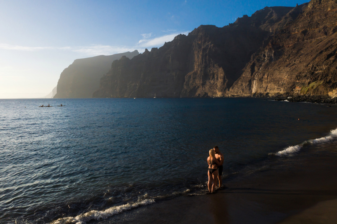 Couple standing at the water&rsquo;s edge beneath the towering Los Gigantes cliffs on Tenerife, with the dark volcanic shoreline and Atlantic Ocean illuminated by evening light.