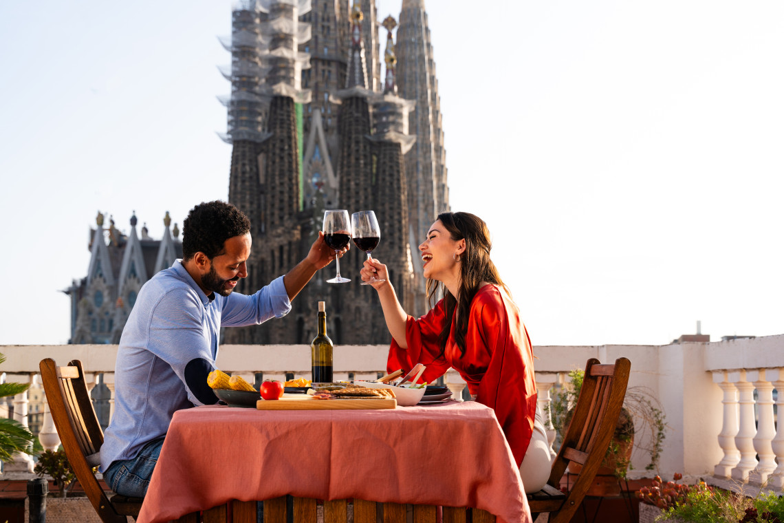 Couple enjoying wine and a meal on a rooftop terrace in Barcelona with the towers of the Sagrada Fam&iacute;lia rising directly behind them.