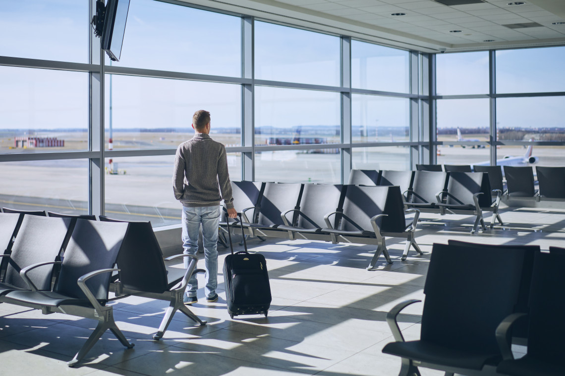 Male traveler standing with suitcase at an empty airport departure gate looking out at the tarmac through floor-to-ceiling windows