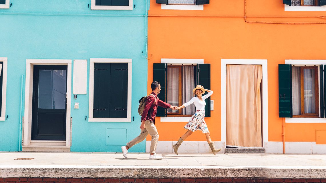 Couple holding hands and walking past vibrant turquoise and orange painted houses on Burano Island, Venice, Italy