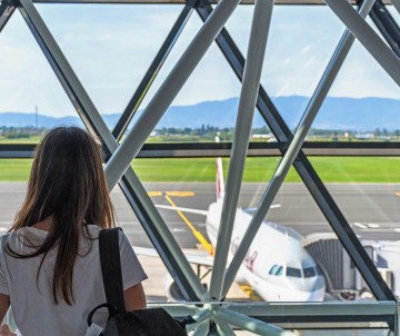 Woman with backpack looking through airport terminal window at plane on the tarmac, preparing to travel to Europe