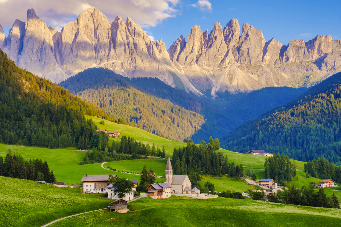 Panoramic view of Santa Maddalena village with a stone church surrounded by green meadows and alpine farmhouses below the dramatic Odle rocky peaks, Val di Funes, South Tyrol, Dolomites, Italy