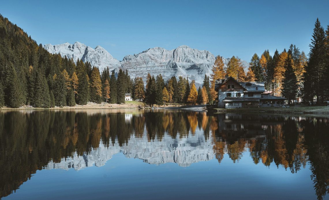 Perfect mirror reflection of autumn larch trees, mountain refuge and Dolomite peaks in the still waters of Lago di Nambino, Madonna di Campiglio, Trentino, Italy