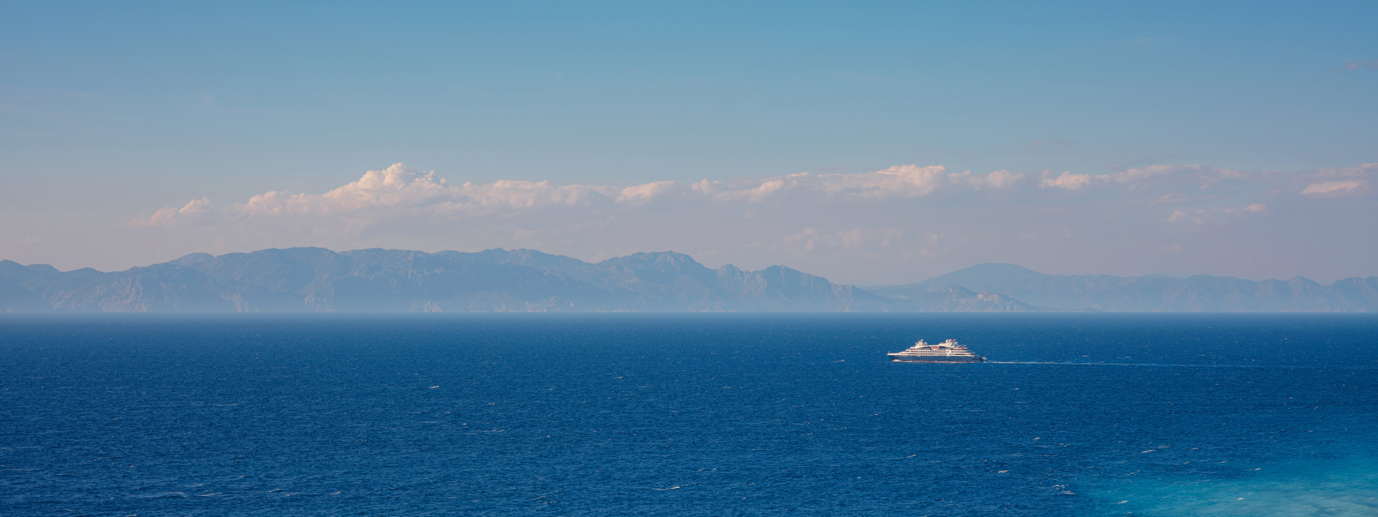 Cruise ship sailing across the deep blue Aegean Sea in Greece, with the mountainous coastline of a Greek island stretching across the horizon.