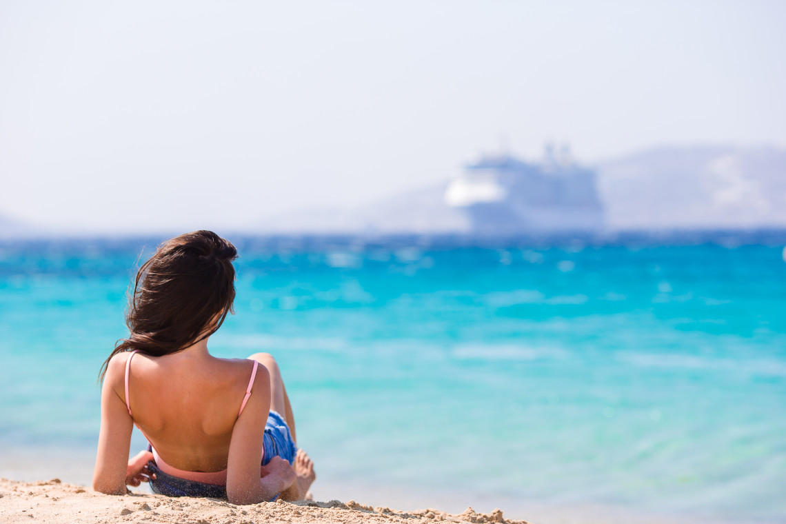 Woman relaxing on a sandy beach beside clear turquoise water in the Greek Islands, with a cruise ship visible offshore in the Aegean Sea.